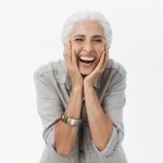 Can Dentures Offer a Natural Smile Transformation for Women? Life only starts when get older. Portrait of charming happy and carefree european senior woman with grey hair laughing out loud from jou and amusement holding palms on cheeks bending from laughter