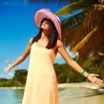 Best Caribbean Holiday Ideas for Girl Bosses beautiful happy woman in colorful sunhat and dress walking near beach ocean on hot summer day near palm