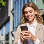 Beyond the Bank: How FinTechs Redefined Cross-Border Payroll Infrastructure Portrait of stylish businesswoman in corporate outfit, holding smartphone, standing on street and using mobile phone in hands